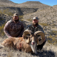 Two men in camouflage pose outdoors with a large ram they have hunted, against a backdrop of dry, hilly terrain.