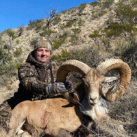 A person in camouflage clothing poses with a large ram in a dry, rocky landscape under a clear sky.