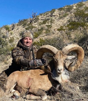 A person in camouflage clothing poses with a large ram in a dry, rocky landscape under a clear sky.