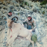 Two men in camouflage pose with a large, horned ram lying on rocks in a dry, grassy landscape.