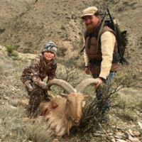 An adult and child pose with a hunted ram on a rocky hillside in a dry, mountainous landscape.