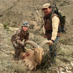 An adult and child pose with a hunted ram on a rocky hillside in a dry, mountainous landscape.