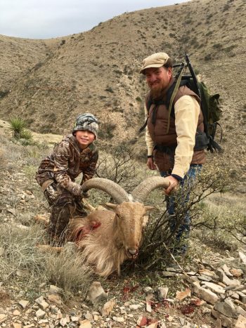 An adult and child pose with a hunted ram on a rocky hillside in a dry, mountainous landscape.