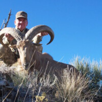 Person in camouflage poses behind a large wild ram with curved horns on a grassy hillside under a clear blue sky.