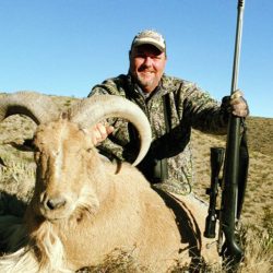 ???????????????????????????????????????? A man in camouflage holds a rifle and poses with a large, horned sheep in a dry, hilly landscape.