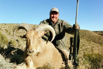 A man in camouflage holds a rifle and poses with a large, horned sheep in a dry, hilly landscape.