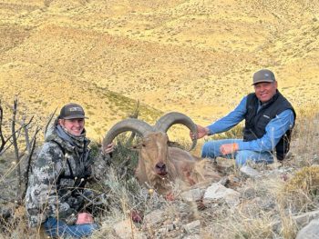 Two men pose with a hunted ram lying on rocky terrain in a dry, hilly landscape.