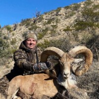 A person in camouflage poses outdoors with a large, horned ram on rocky terrain under a clear blue sky.